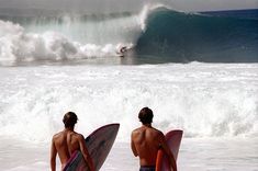 two surfers are standing in the water with their surfboards looking at an ocean wave