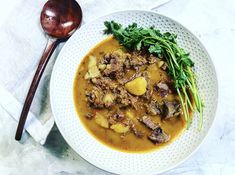 a white bowl filled with stew and greens next to a spoon on top of a table