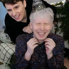 an older man wearing a birthday hat and smiling at the camera while another person holds his neck around him