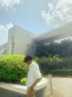 a man standing next to a white wall and green bushes in front of a building