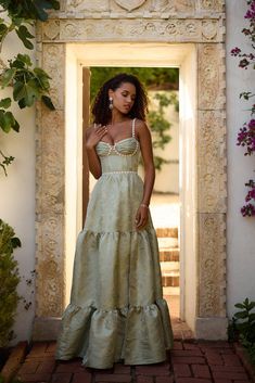 a woman in a long dress is standing by an archway with flowers on the side