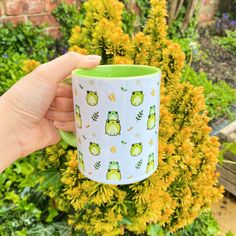 a hand holding a green and white coffee mug in front of some yellow flower bushes