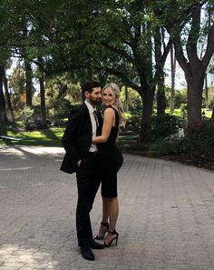 a man and woman standing next to each other on a brick walkway with trees in the background