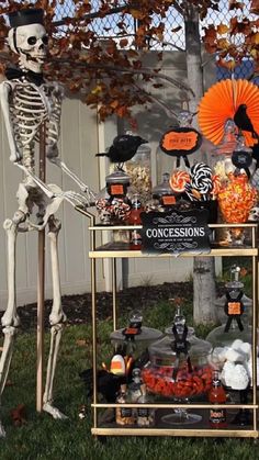 a skeleton sitting on top of a table filled with halloween decorations and candies in front of a fence