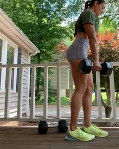 a woman in grey shorts and green shirt holding two black dumbbells while standing on a porch