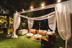 an outdoor area with couches, tables and white drapes on the grass at night