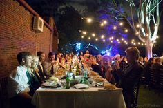 a group of people sitting around a table eating food and drinking wine at night time