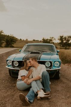 a man and woman sitting on the ground next to a blue mustang in front of a car