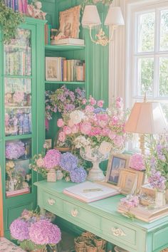 a green desk topped with lots of flowers next to a book shelf filled with books