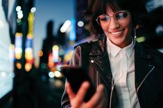 a woman wearing glasses is looking at her cell phone in the city with headphones on