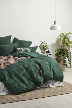 a bed with green comforter and pillows in a white room next to potted plants