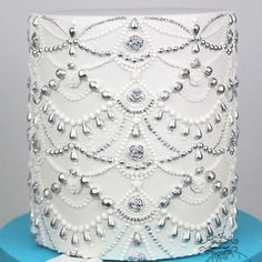 a white wedding cake with silver beads and pearls on the top is sitting on a blue stand