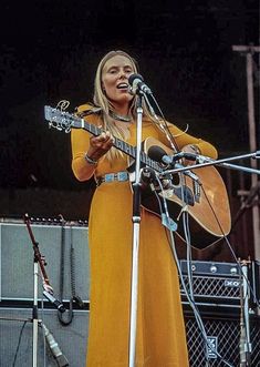 a woman singing into a microphone while holding an acoustic guitar in front of a stage