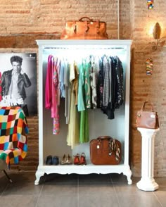 a white cabinet with clothes hanging on it and a suitcase sitting next to it in front of a brick wall