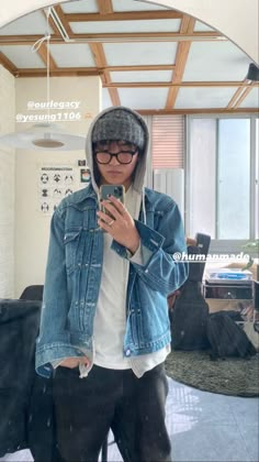 a man wearing glasses and a denim jacket is looking at his cell phone while standing in front of a mirror