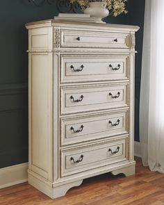 an antique white dresser in a room with dark walls and wood flooring, along with a vase filled with flowers