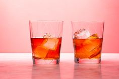 two glasses filled with ice sitting on top of a pink counter next to each other