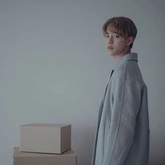 a young man standing next to boxes in front of a white wall and wearing a gray coat