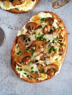 two pizzas with mushrooms and cheese on them sitting on a table next to spoons