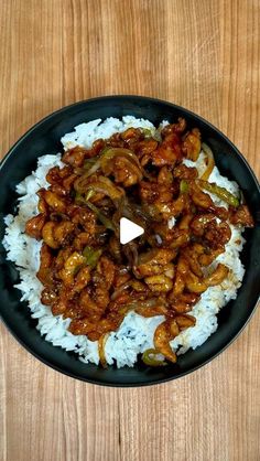 a black plate topped with rice covered in meat and sauce on top of a wooden table