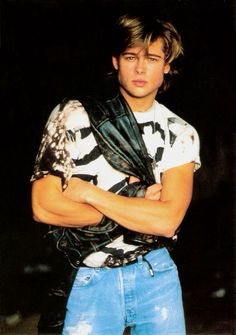 a young man with his arms crossed wearing a leather vest and ripped denims, standing in front of a black background