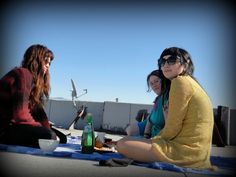 three women sitting on the ground with food and drinks in front of them, all looking at one another