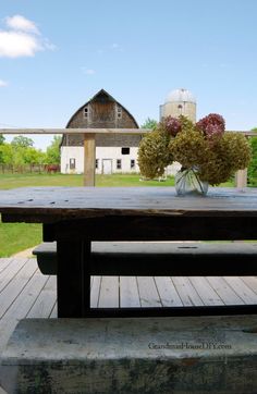 a wooden table sitting on top of a lush green field next to a white barn