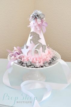 a pink and silver birthday hat sitting on top of a white cake plate with ribbons