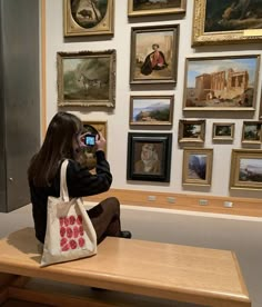 a woman sitting on a bench taking a photo with her cell phone in front of some paintings