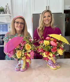 two women standing next to each other with flowers in vases on the counter top