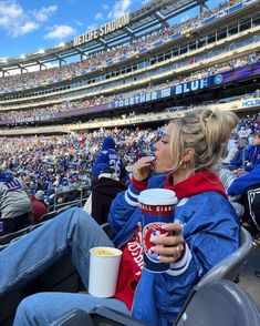 a woman sitting in the stands at a football game eating popcorn and drinking from a cup