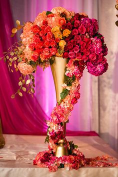 a tall gold vase filled with lots of colorful flowers on top of a white table