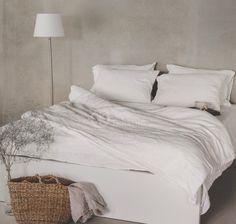 a bed with white sheets and pillows next to a basket on the floor near a lamp