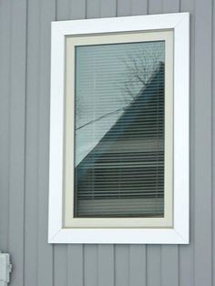 a window with blinds on the side of a gray building and a tree in the reflection