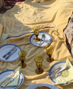 a table set with plates and silverware on a yellow blanket in the sand at the beach