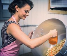 a woman pouring orange juice into a large metal pot filled with popcorn and corn kernels