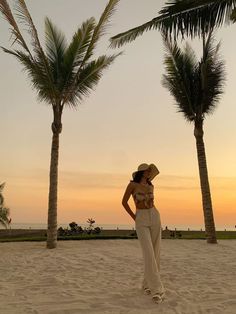 a woman standing on top of a sandy beach next to two palm trees at sunset