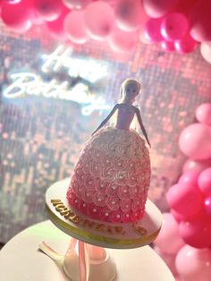 a barbie doll on top of a pink cake with balloons in the backgroud