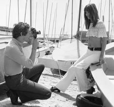 a man kneeling down next to a woman with a camera in front of sailboats