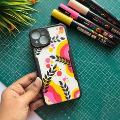 a person holding an iphone case on top of a table next to markers and pencils