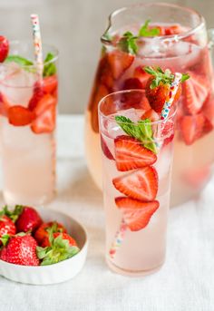 two glasses filled with water and strawberries on top of a white table next to bowls of strawberries