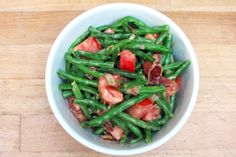 a white bowl filled with green beans, bacon and tomatoes on top of a wooden table