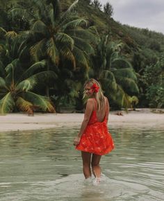 a woman in a red dress is standing in the water with palm trees behind her