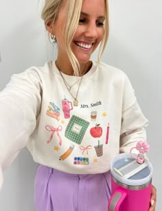 a woman holding a pink coffee mug and wearing a white sweatshirt with school items on it