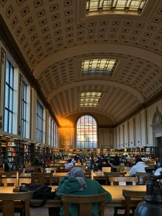 people are sitting at tables and reading books in a large room with high ceilinged windows