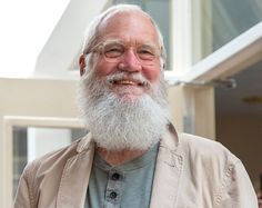 an older man with a long white beard and glasses smiles at the camera while standing in front of a building