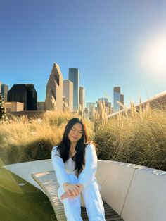 a woman sitting on top of a wooden bench in front of tall grass and buildings