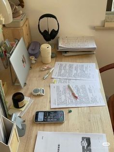 a wooden desk topped with lots of papers and headphones next to a phone on top of it