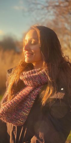 a woman with long hair wearing a scarf and smiling at the camera while standing in a field