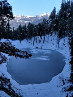 a frozen pond surrounded by trees in the middle of snow covered ground with mountains in the background